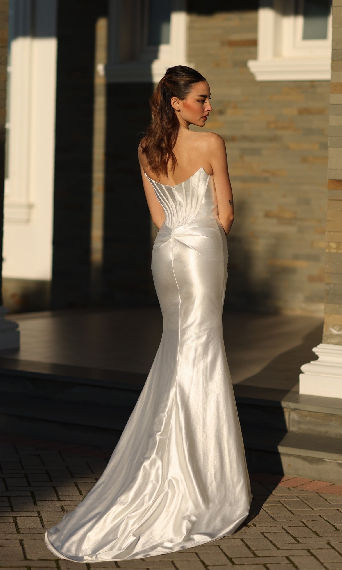 A woman in the Strapless Off-White Formal Dress: Tina Holly TK151W stands on a stone patio outdoors, facing away from the camera toward a building with stone walls and white trim.